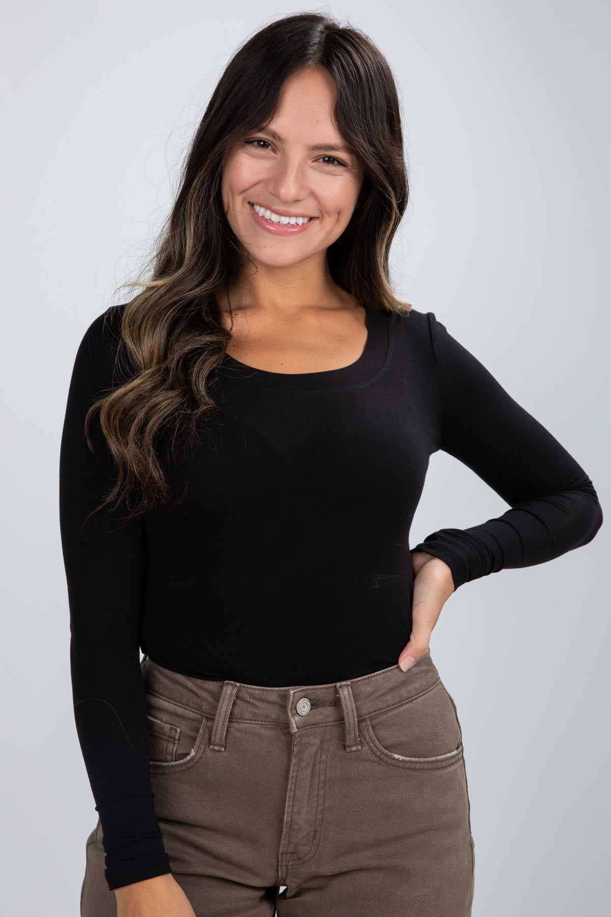 Woman wearing a black long-sleeve top and brown pants against a gray background