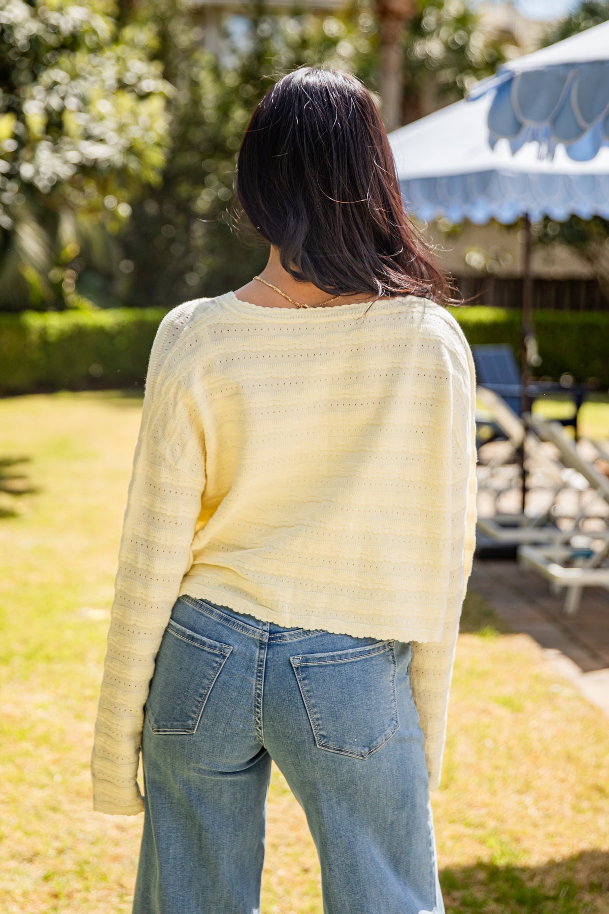 Person wearing a light yellow sweater and blue jeans standing outdoors with greenery and a tent in the background.