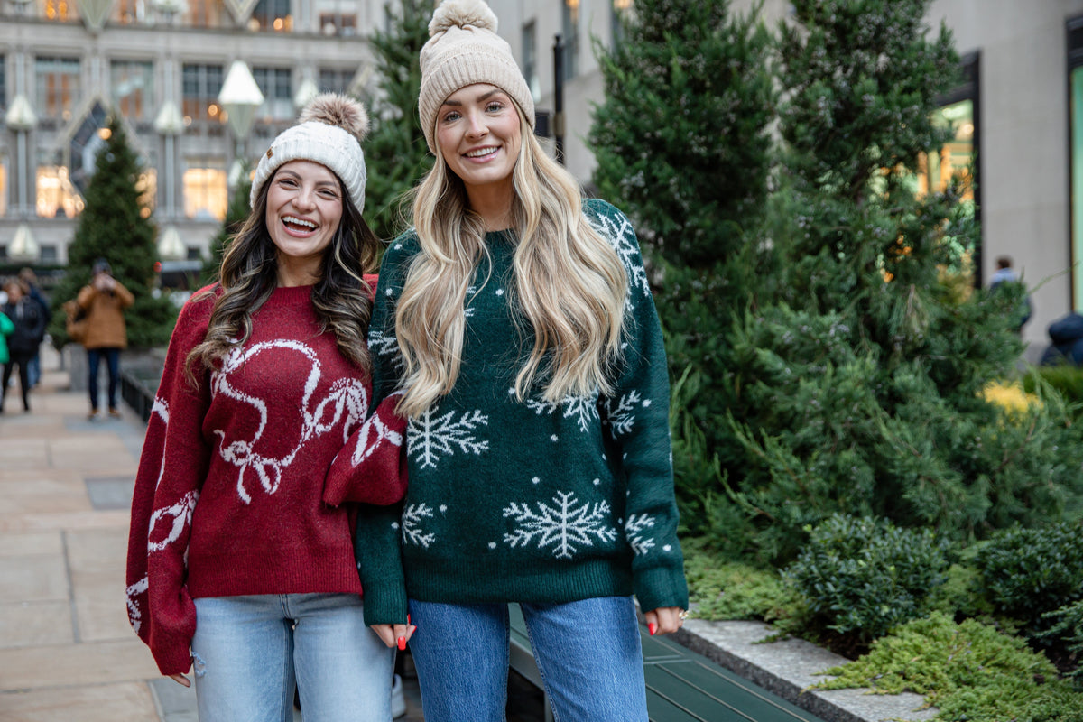 Two women smiling outdoors near evergreen trees, wearing winter knit sweaters and beanies. One wears a red sweater with a white holiday graphic and light-wash jeans, while the other wears a dark green sweater with white snowflake details and blue jeans.