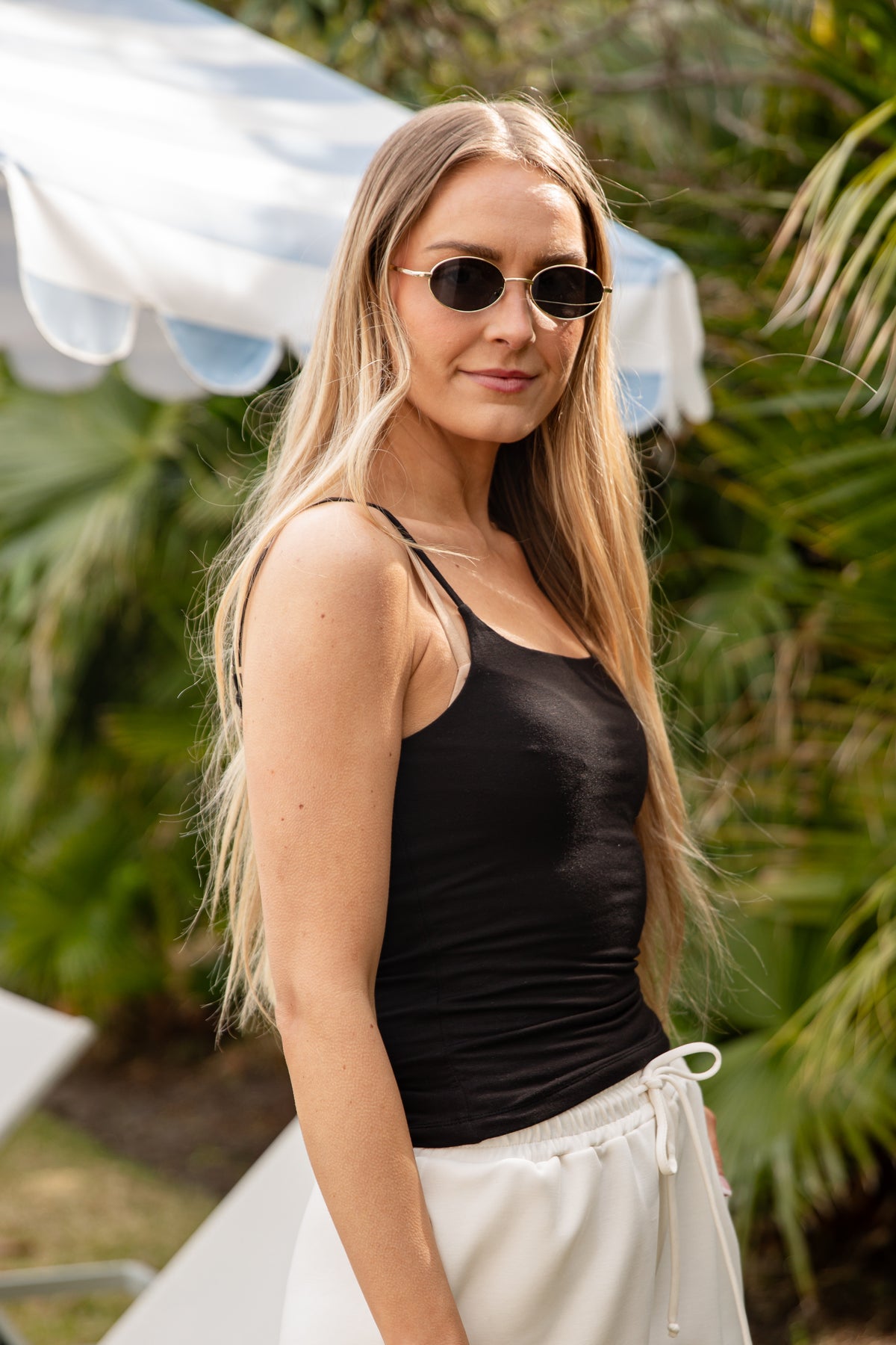 Woman wearing sunglasses and a black tank top with white pants outdoors.
