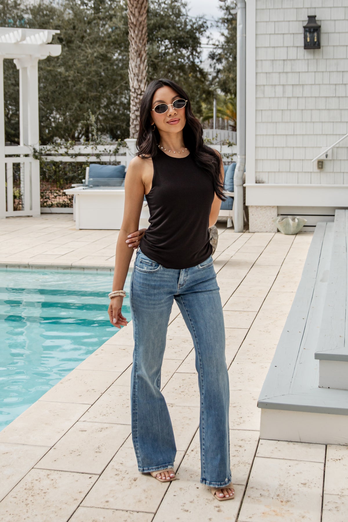Woman in black tank top and blue jeans standing by a poolside.