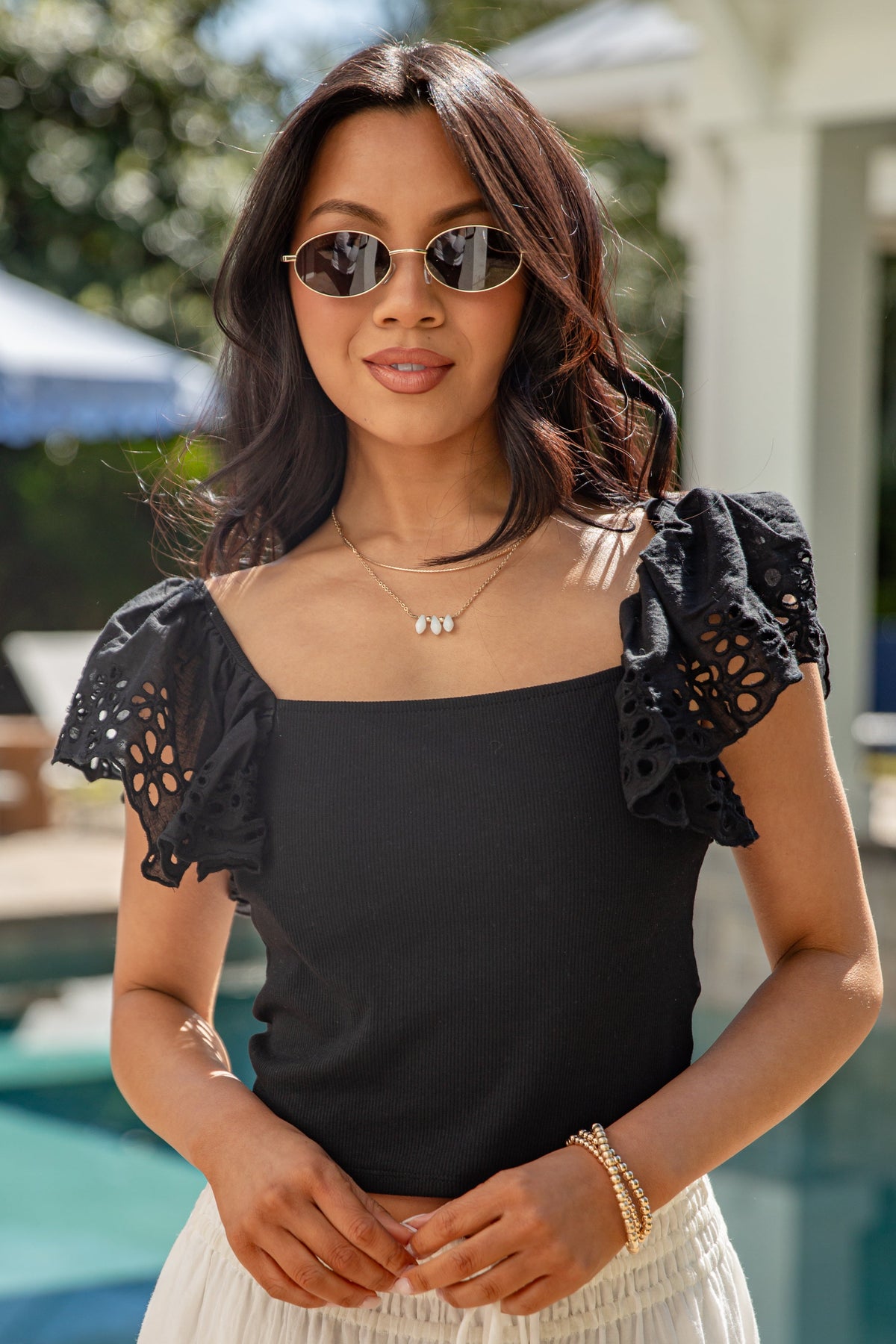 Woman in black eyelet-sleeve top and cream skirt by pool.