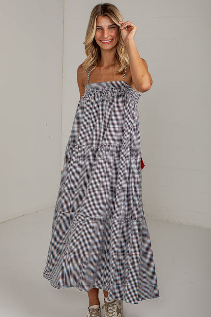 A woman modeling a sleeveless, tiered maxi dress with a small black and white gingham pattern, paired with beige sneakers, in a bright studio setting.