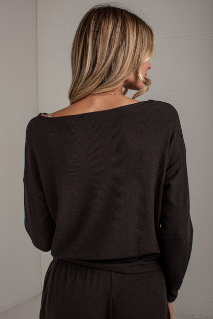 A woman viewed from behind wearing a dark brown off-the-shoulder knit sweater and matching dark bottoms, standing against a light gray paneled wall.