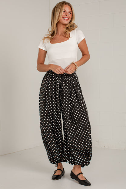 A woman modeling black wide-leg pants with a white polka dot pattern, paired with a white scoop-neck t-shirt and black Mary Jane shoes, standing against a white brick wall.