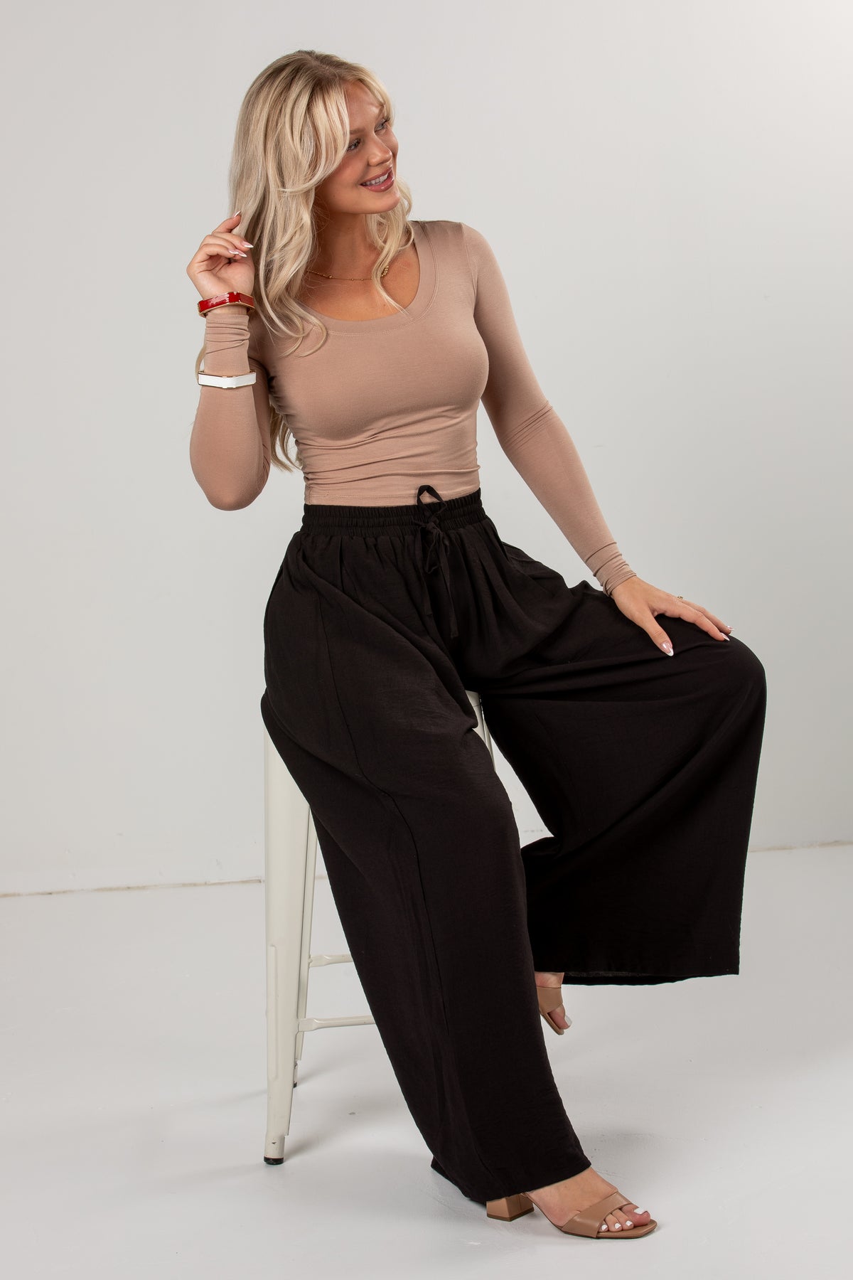 A woman models a beige long-sleeve scoop-neck top and black wide-leg drawstring pants, sitting on a white stool against a light gray studio background.