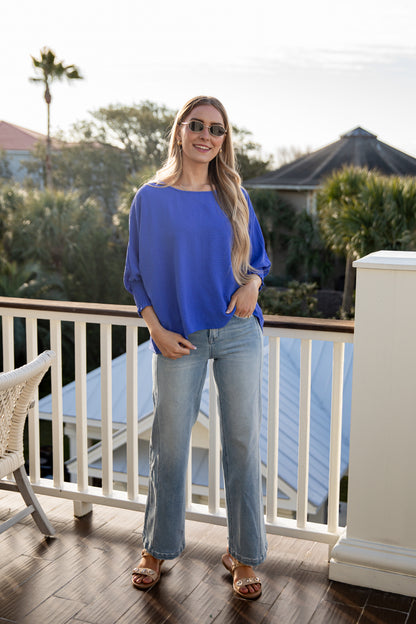 Woman in a blue shirt and jeans standing on a wooden deck with greenery in the background