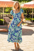 Woman in a blue floral dress standing outdoors with lounge chairs and umbrellas in the background.