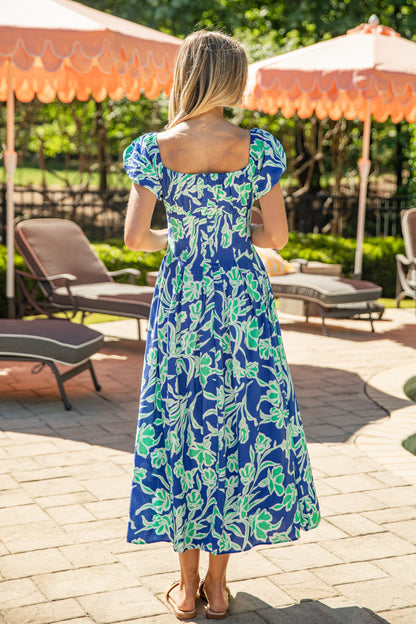 Woman in a blue floral dress standing in a sunlit outdoor setting with lounge chairs and umbrellas.