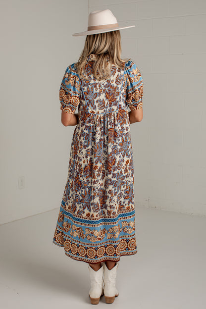 A woman viewed from behind wearing a bohemian-style midi dress with a cream base and blue, brown, and orange floral patterns. She is wearing a wide-brimmed beige hat and white cowboy boots against a white studio background.
