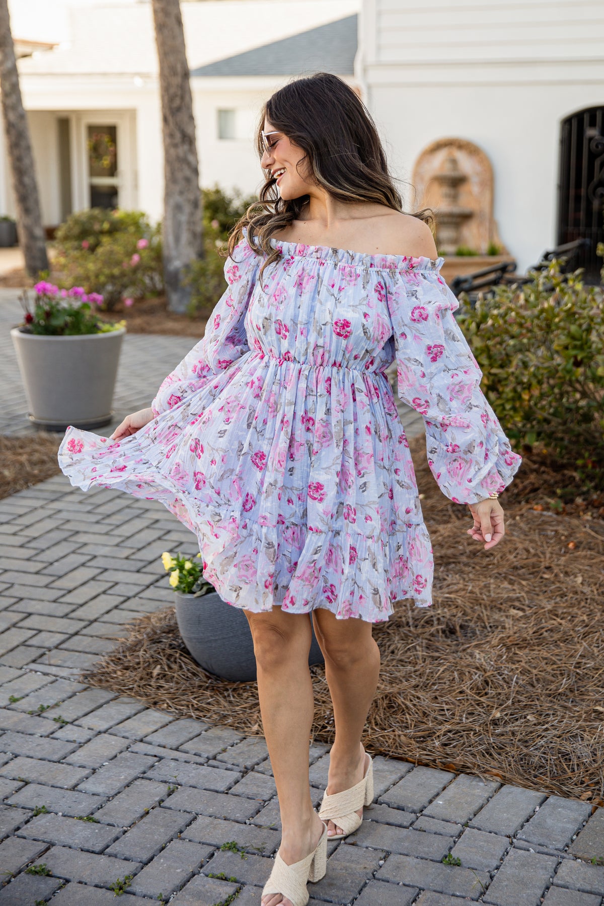 Woman in a floral off-shoulder dress standing on a patio with plants and a house in the background.