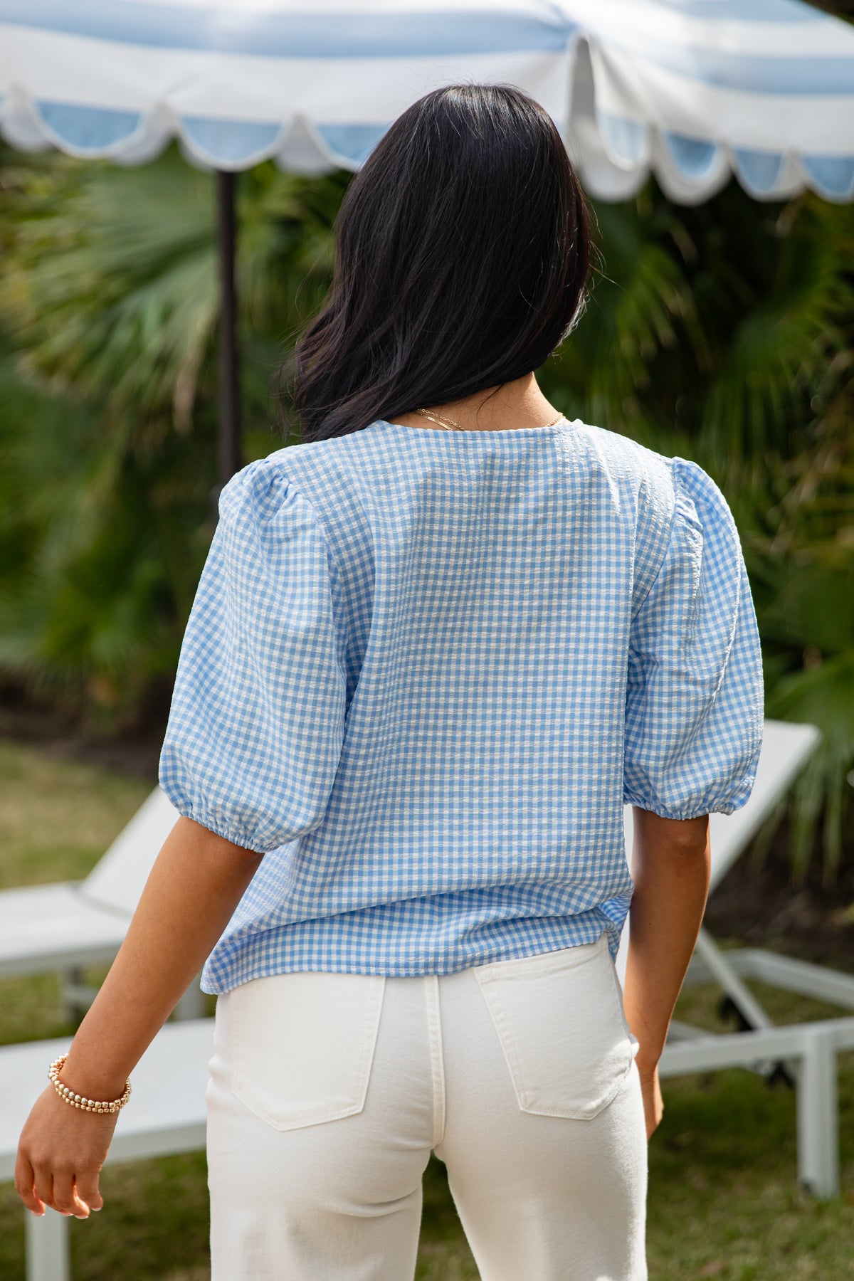 Person wearing a blue checkered shirt and white pants under a white canopy with greenery in the background