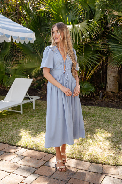 Woman in a light blue dress standing outdoors with greenery and a white chair in the background