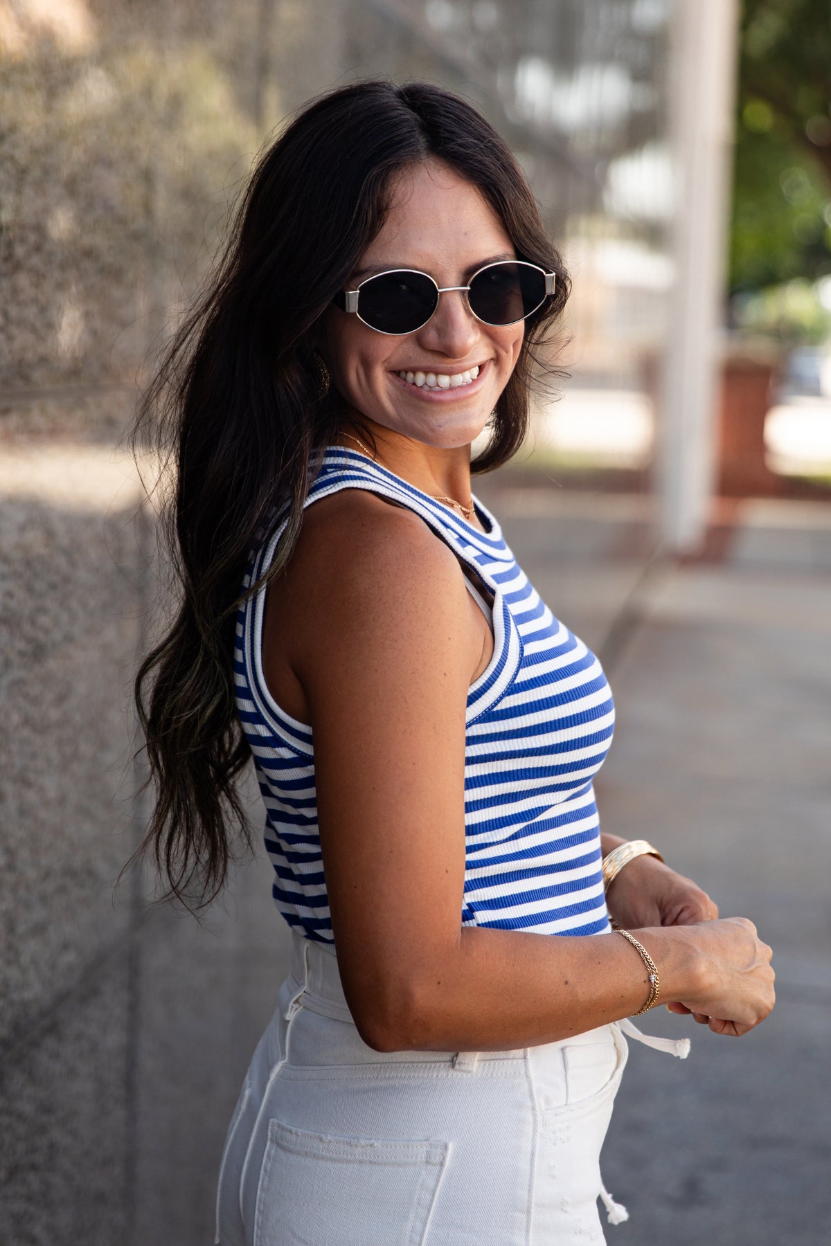 A woman wearing a blue and white striped tank top and white jeans, smiling and looking back over her shoulder in an outdoor setting.