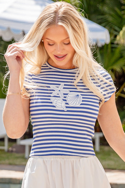 Woman wearing a blue and white striped shirt with a graphic design, standing outdoors near a pool.
