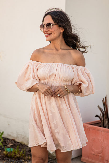 Woman wearing a light pink off-shoulder dress standing outdoors.