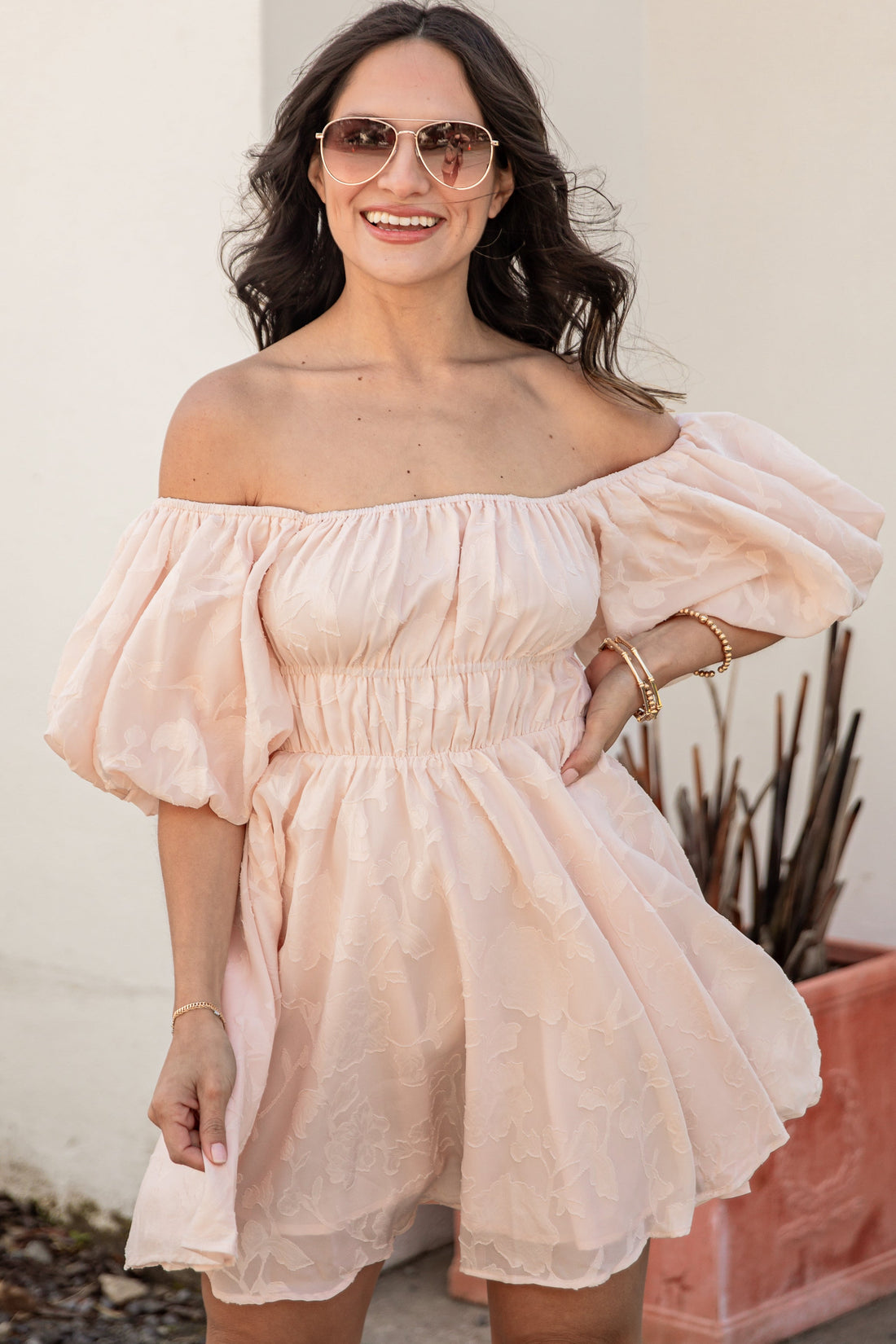 Woman wearing a light pink off-shoulder dress with sunglasses, standing against a white wall.