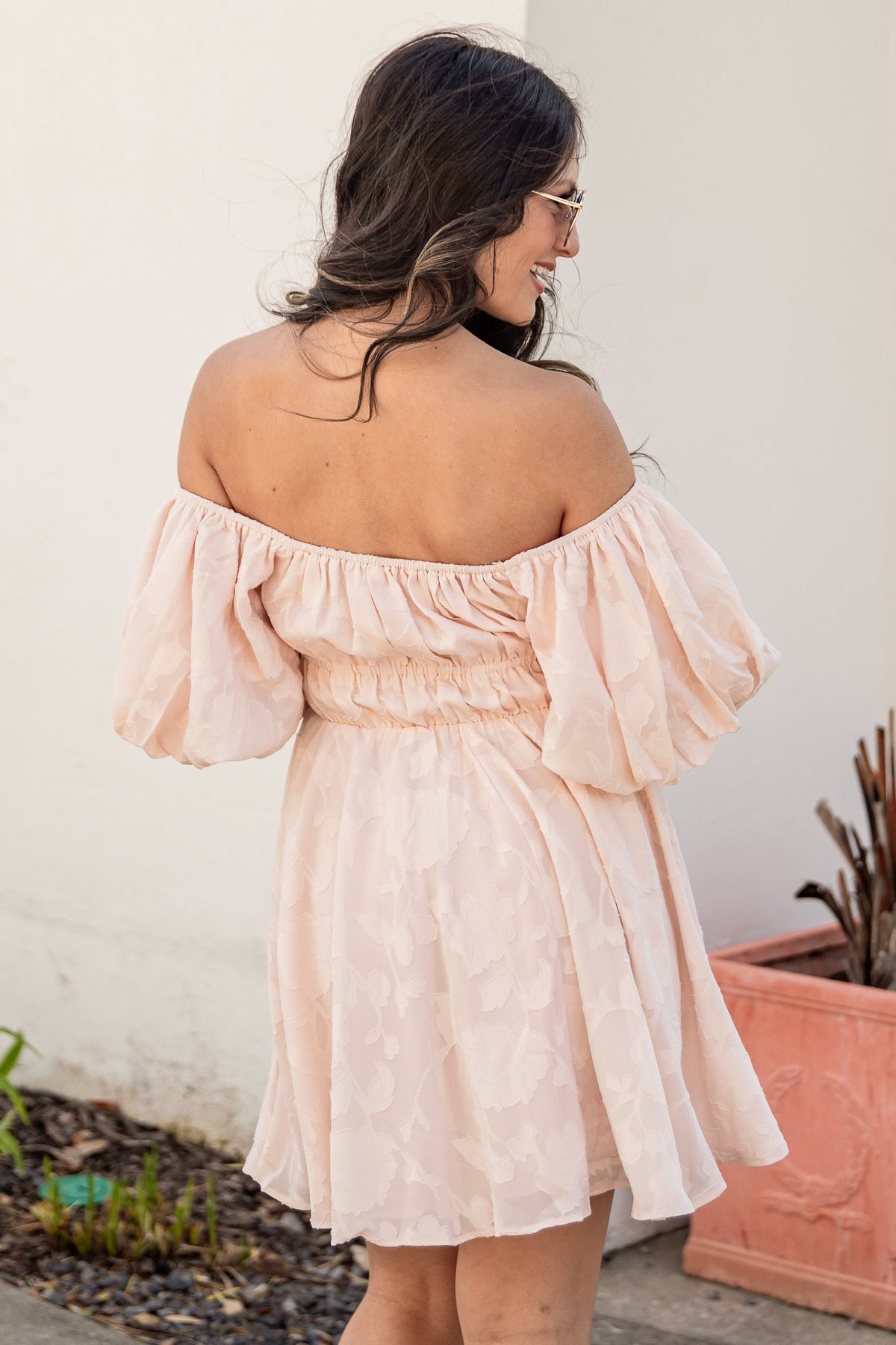 Woman wearing a pink off-shoulder dress standing outdoors next to a plant.