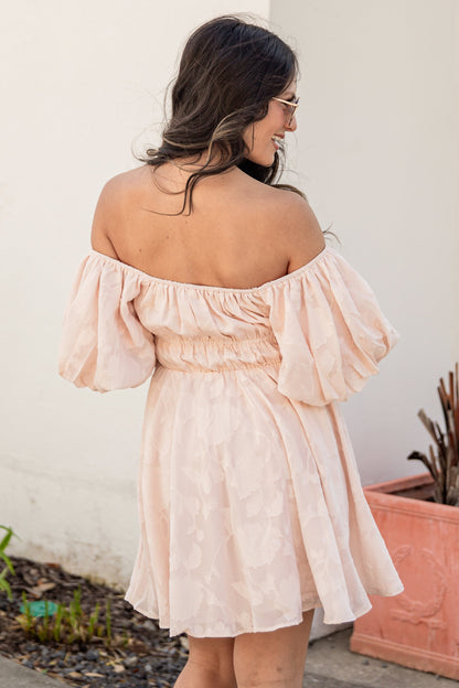 Woman wearing a pink off-shoulder dress standing outdoors next to a plant.
