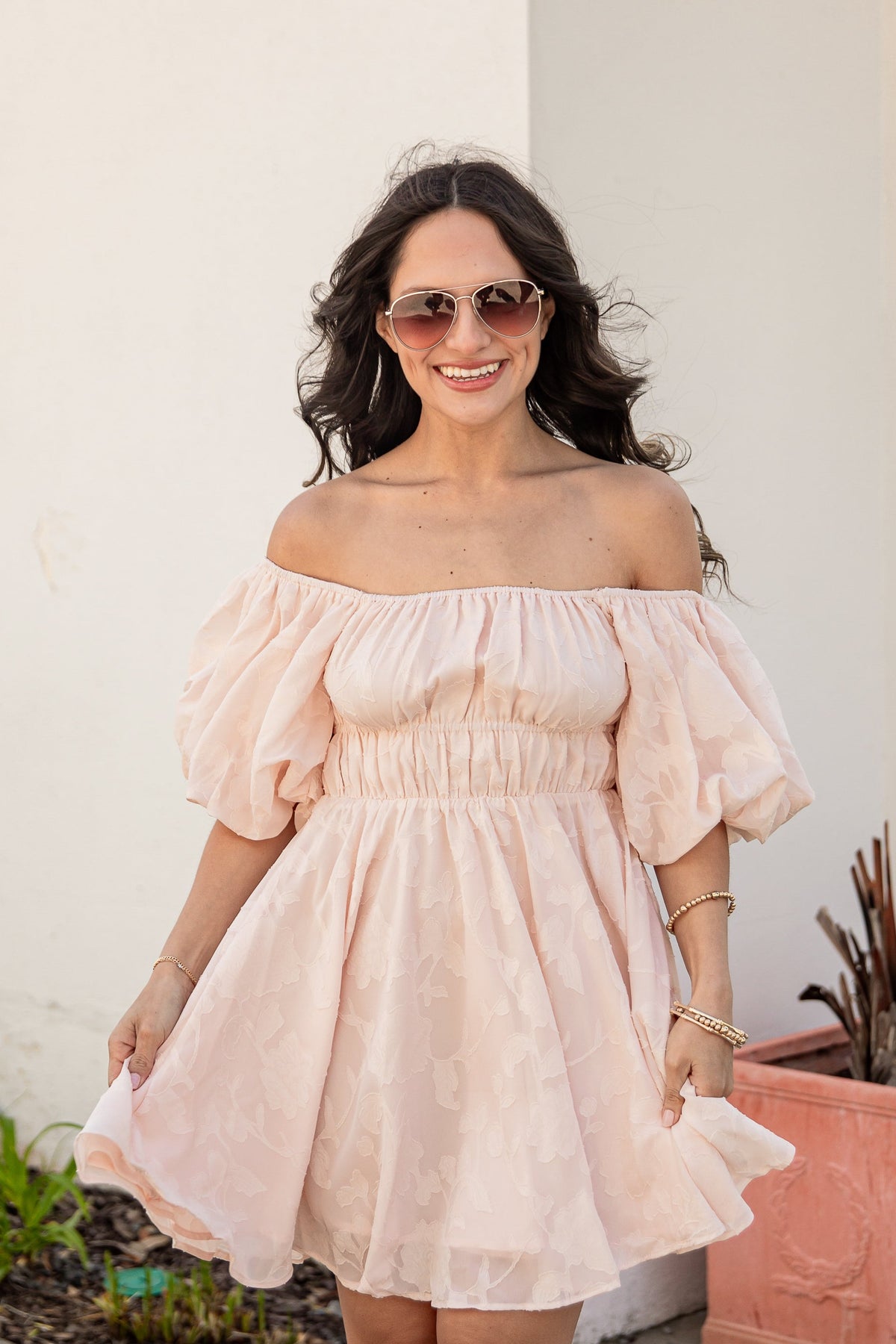 Woman in a light pink off-shoulder dress standing outdoors next to a white wall and potted plant.
