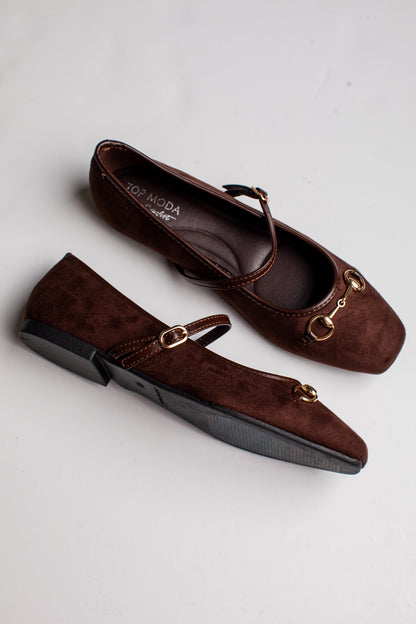 A pair of dark brown suede Mary Jane flats with gold-tone horsebit hardware and a low block heel, displayed on a light gray background.