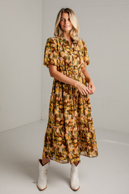 A woman modeling a short-sleeve, tiered midi dress with a warm-toned floral print in yellow, orange, and brown, paired with white cowboy boots against a white studio background.