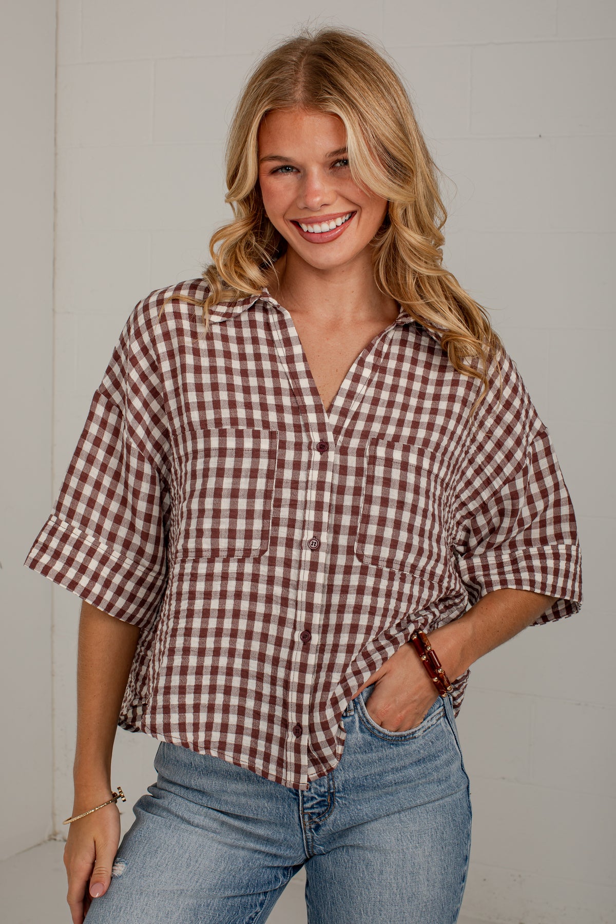 A smiling blonde woman wearing a brown and white gingham check short-sleeve button-up shirt with rolled cuffs and light blue jeans, standing against a light gray wall.