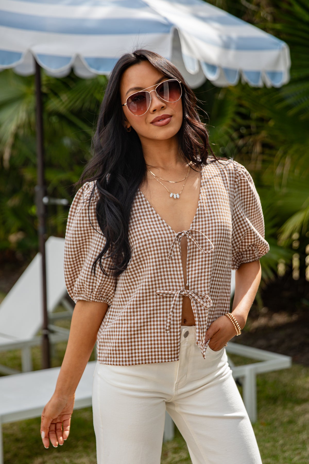 Woman wearing a checkered blouse and sunglasses outdoors with greenery in the background