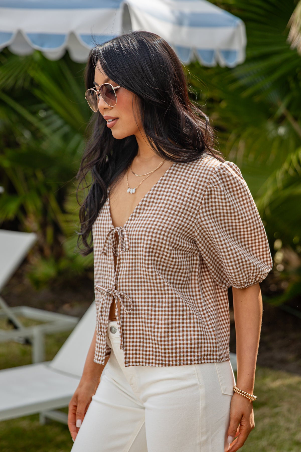Woman wearing a checkered blouse and sunglasses outdoors with greenery in the background