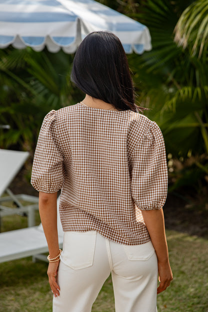 Woman wearing a brown checkered blouse and white pants standing outdoors with greenery and a striped umbrella in the background.