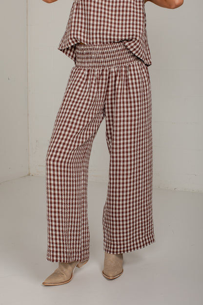 A person wearing brown and beige gingham wide-leg pants with a smocked elastic waist, paired with beige pointed-toe heeled boots, photographed in a plain studio corner.