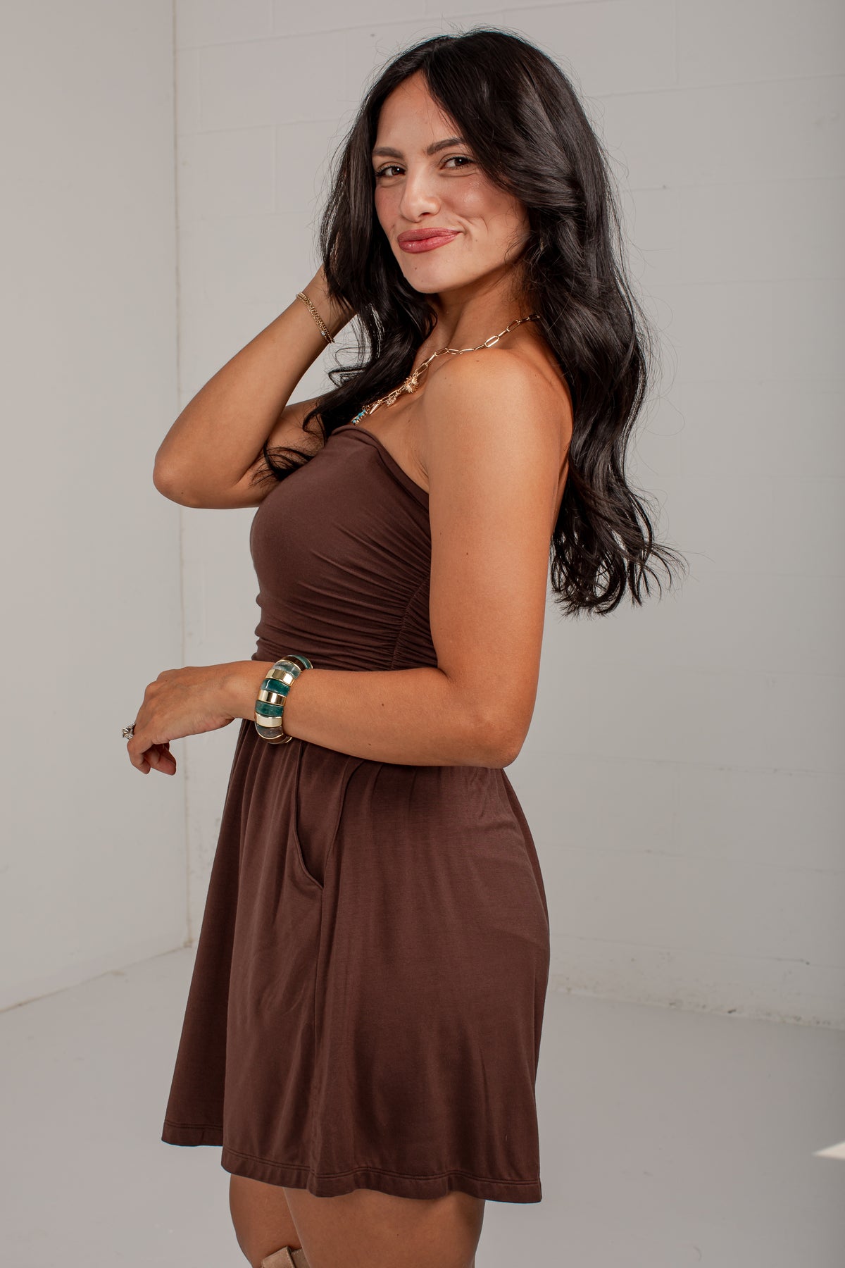 A woman modeling a brown strapless mini dress with a gathered waist and flared skirt, standing against a plain light gray wall.