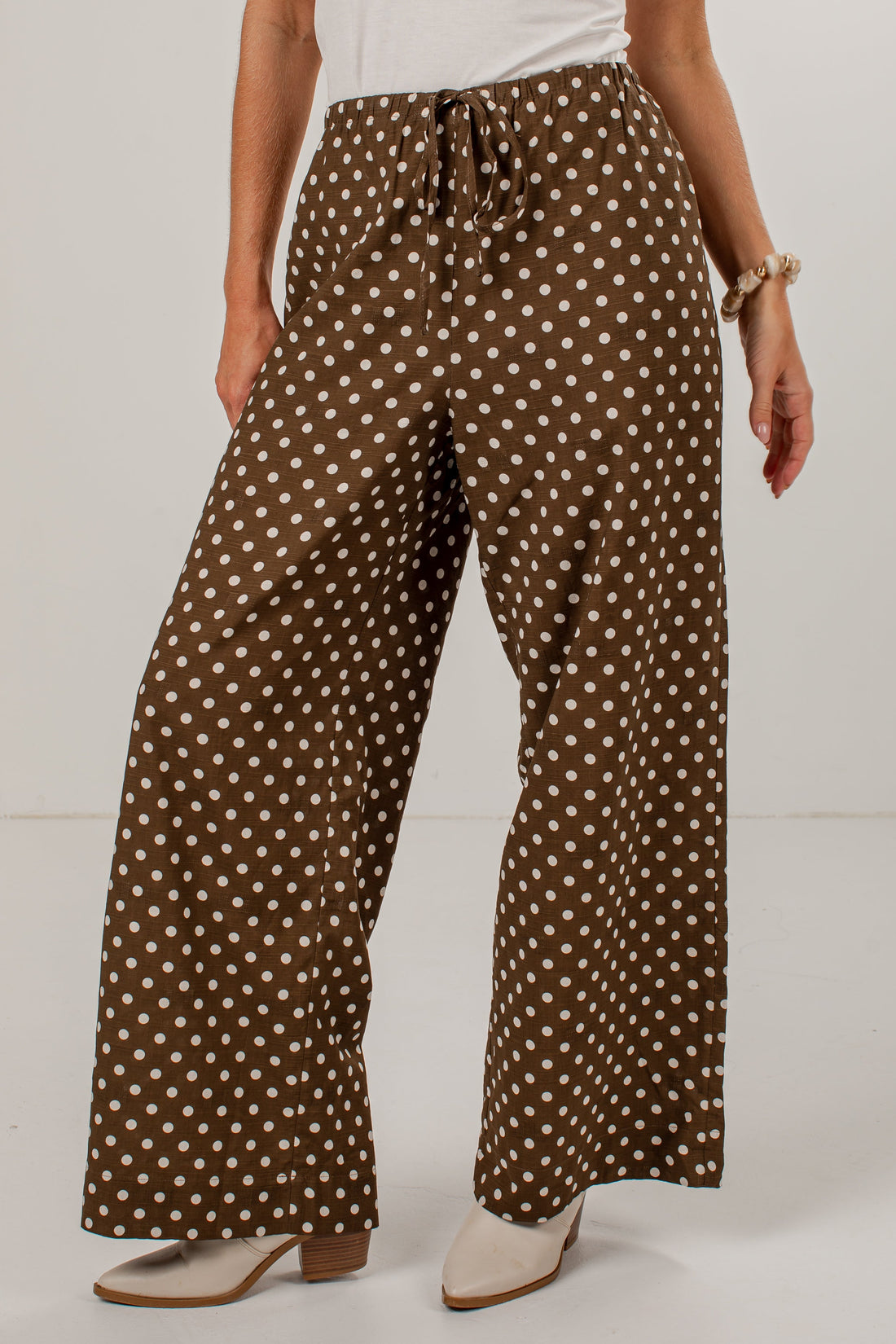 A person wearing brown wide-leg pants with a white polka dot pattern, styled with a white top and beige heeled shoes against a plain white studio background.