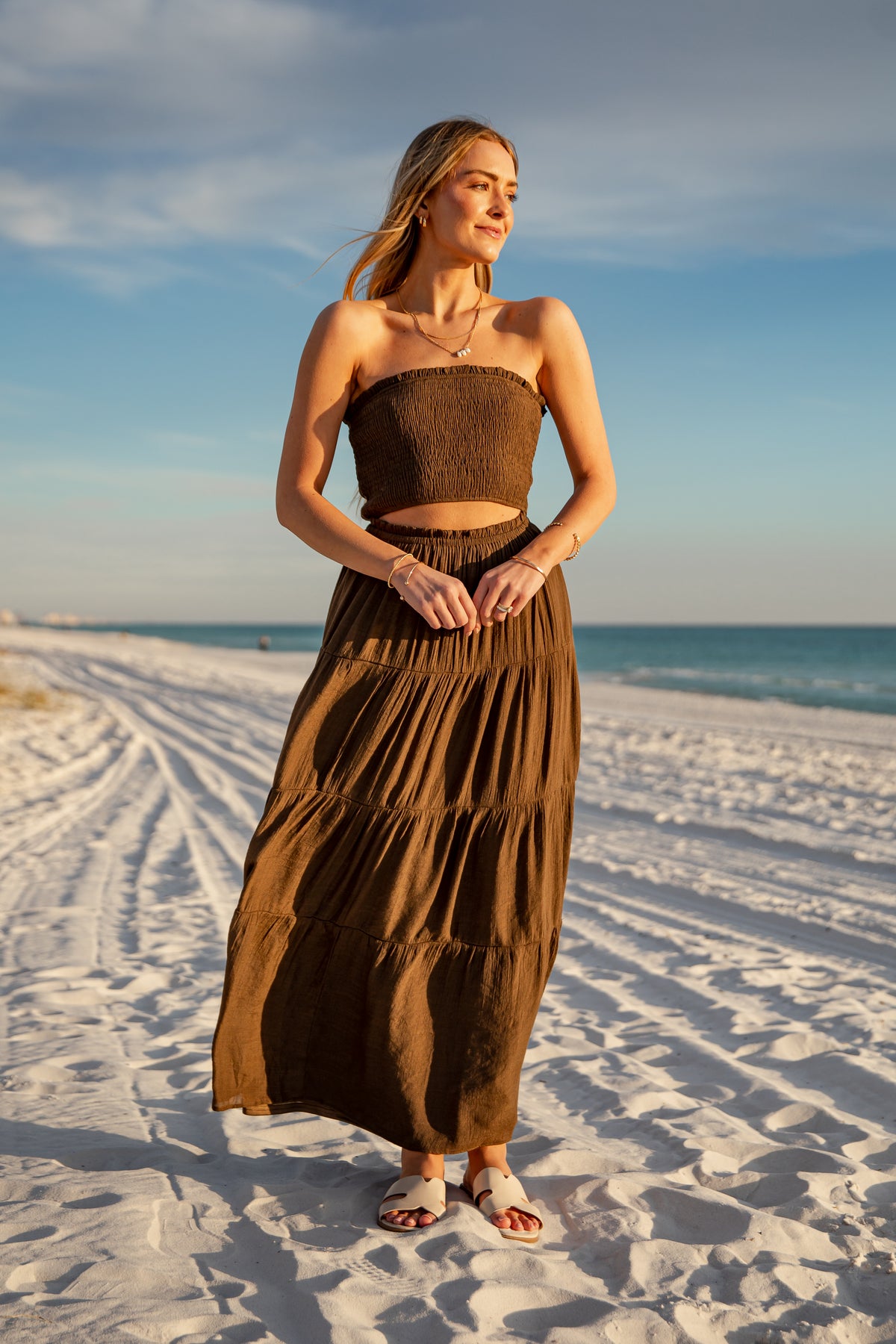 Woman in a brown dress standing on a sandy beach with ocean and sky in the background