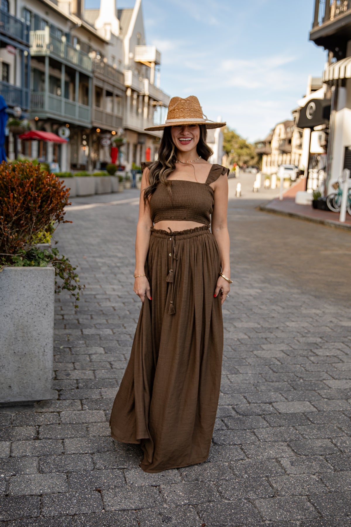 Woman in a brown dress and hat standing on a street with buildings and trees in the background.