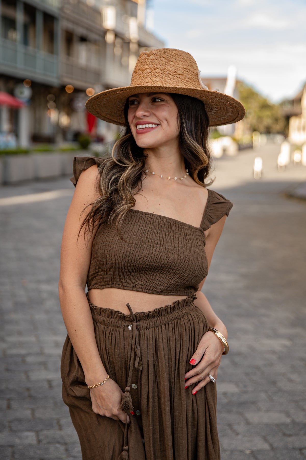 Woman in a brown outfit with a wide-brimmed hat standing on a street.