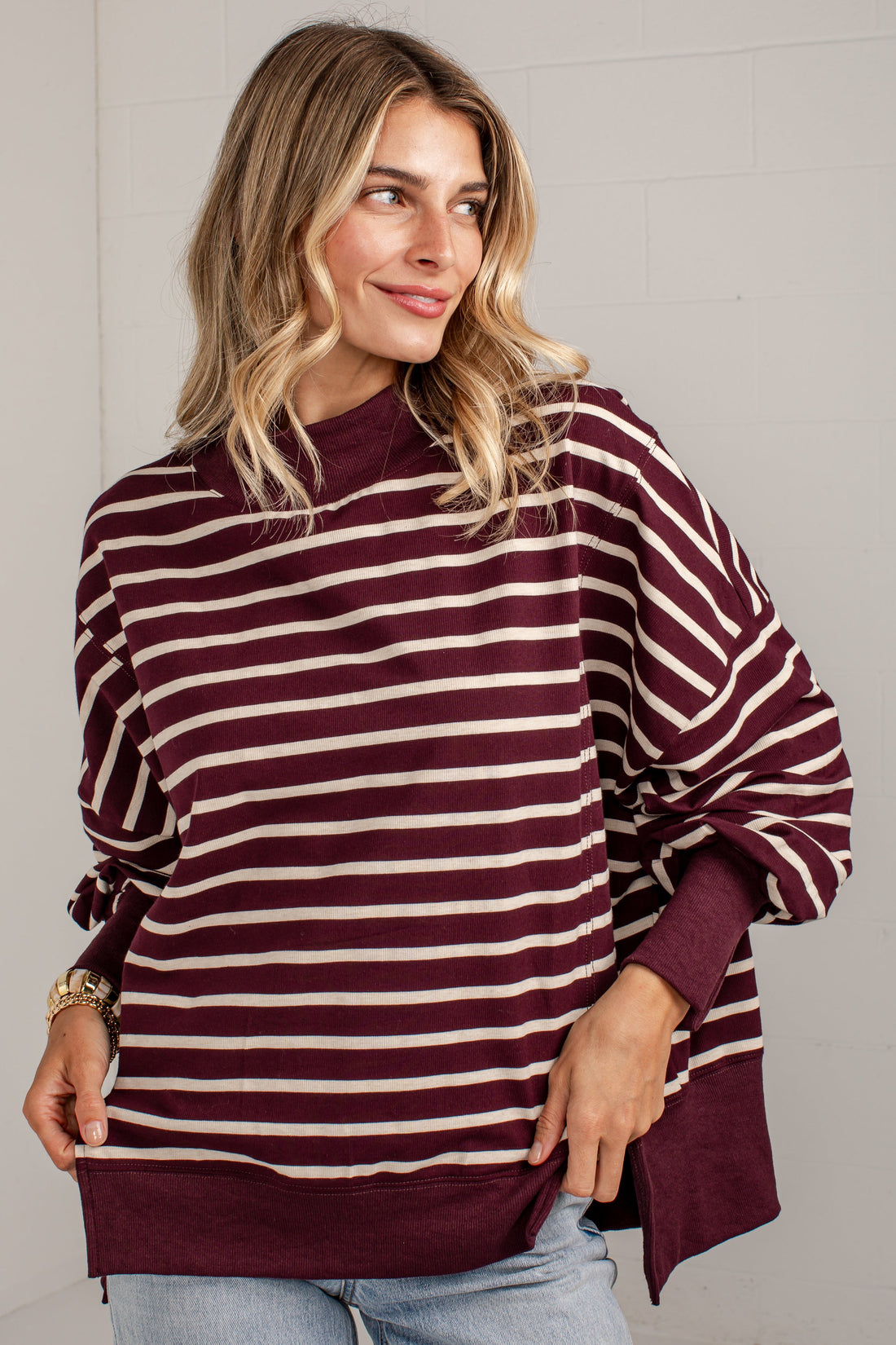 A woman wearing a loose-fitting, long-sleeve striped sweater in burgundy and cream, paired with light-wash jeans, standing against a white brick wall.