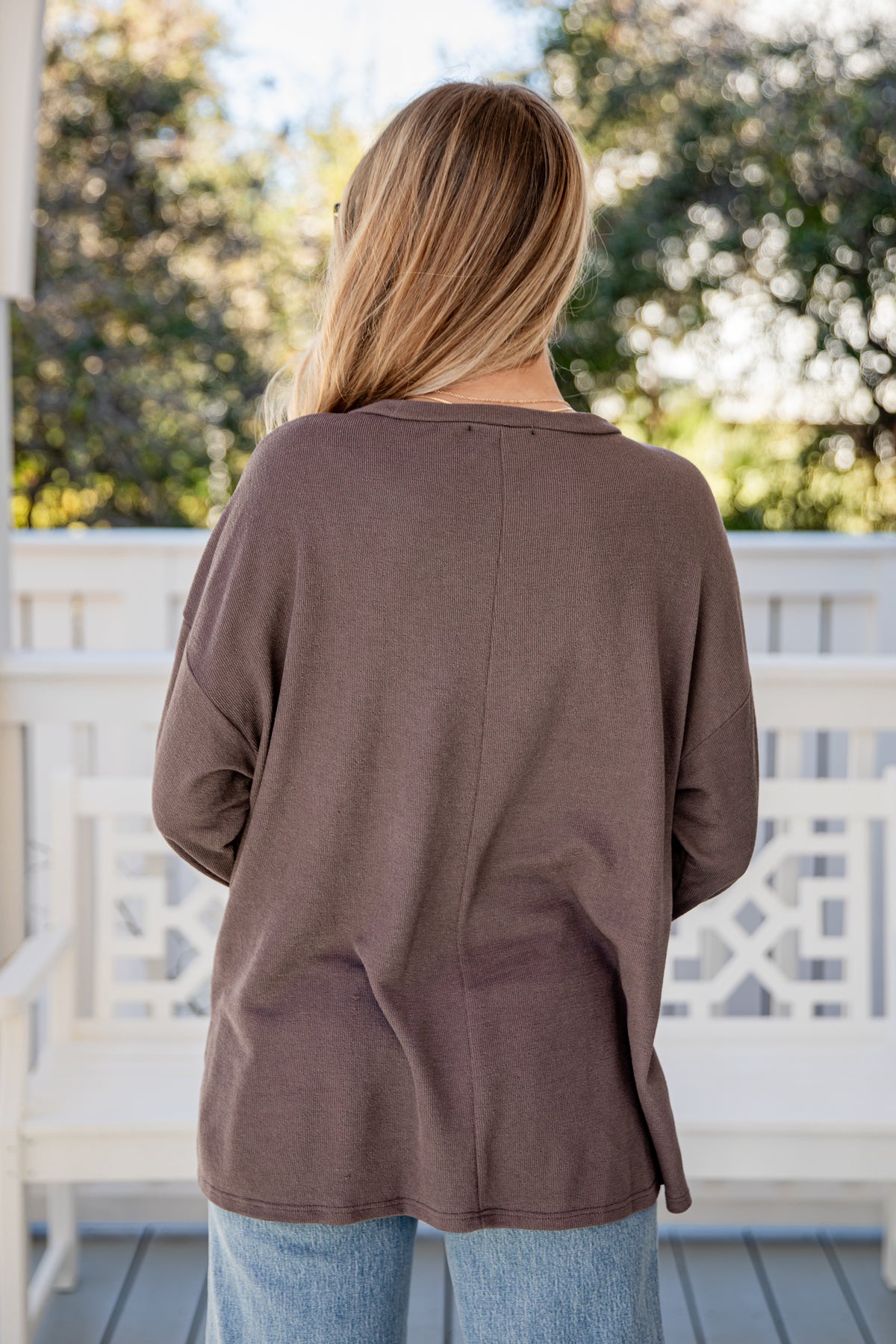 Woman wearing a brown sweater on a porch with blurred greenery in the background
