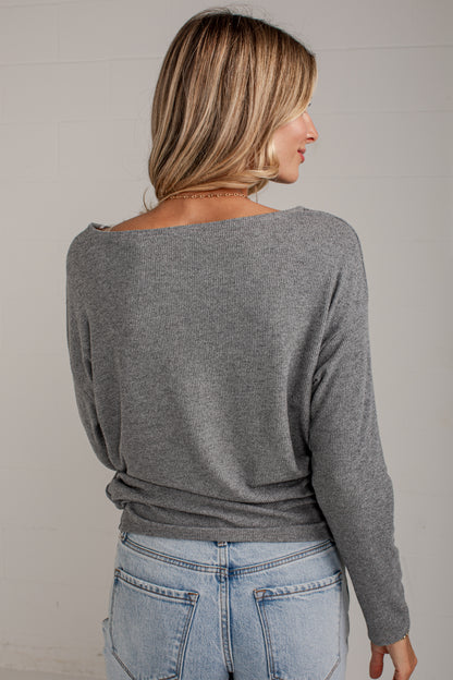 A woman shown from the back wearing a heather gray long-sleeve knit top with a wide boat neckline, paired with light blue jeans, against a light gray wall.