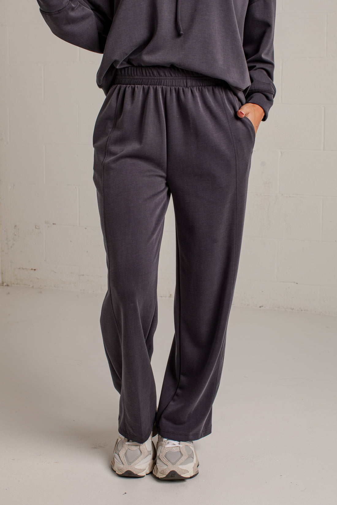 A person wearing dark charcoal gray sweatpants with an elastic waistband and side pockets, paired with a matching dark hoodie and light gray sneakers, standing against a light gray brick wall.