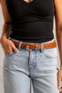 A close-up of a person wearing a slim brown leather belt with a polished gold-tone oval buckle, styled with light-wash blue jeans and a black ribbed tank top.