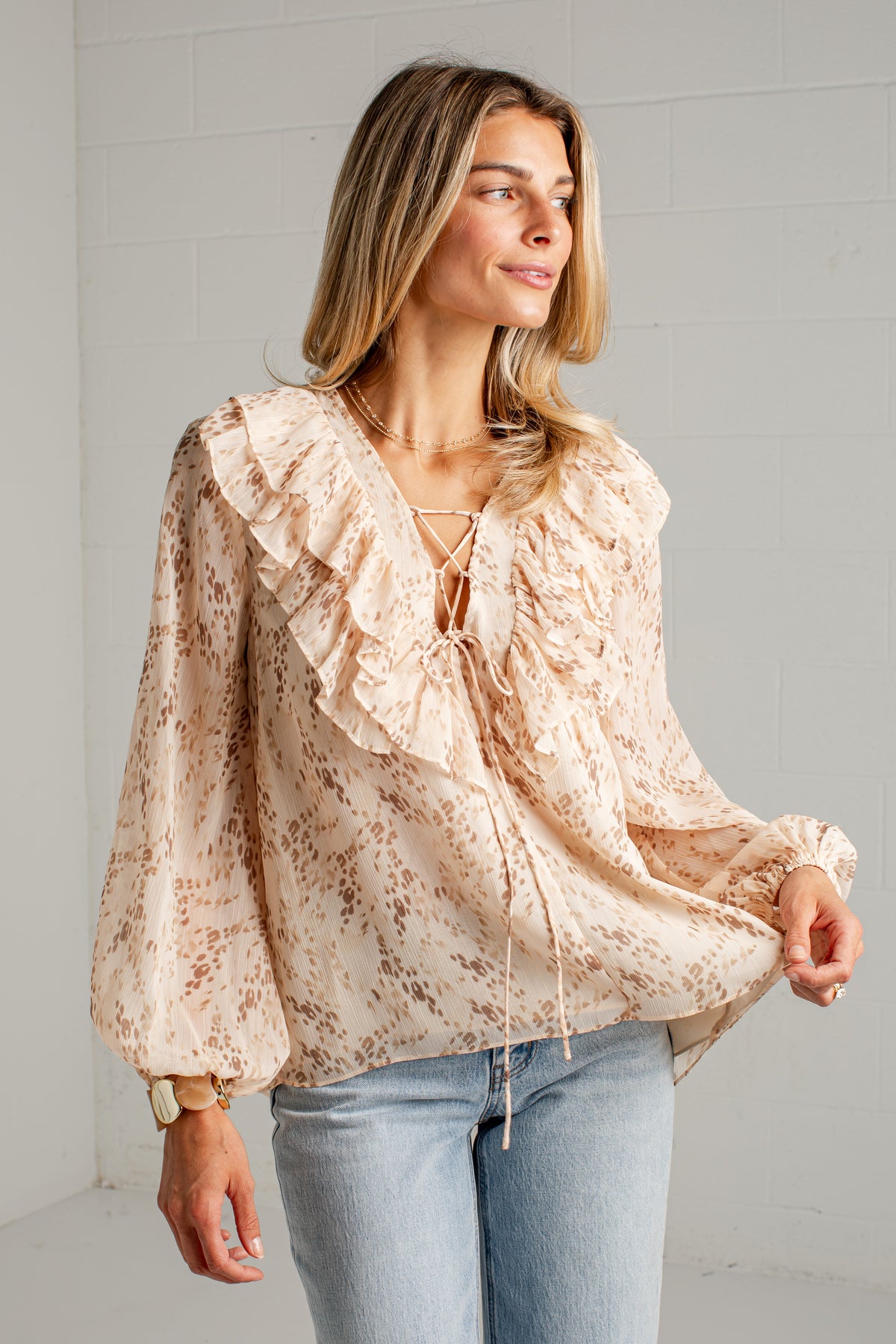 A woman wearing a beige and brown print blouse with ruffles and tie details, paired with light blue jeans, standing against a white brick wall.