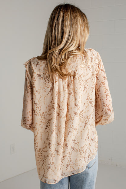 A woman viewed from behind wearing a loose beige blouse with a brown leopard print pattern and light blue jeans, standing against a white wall.