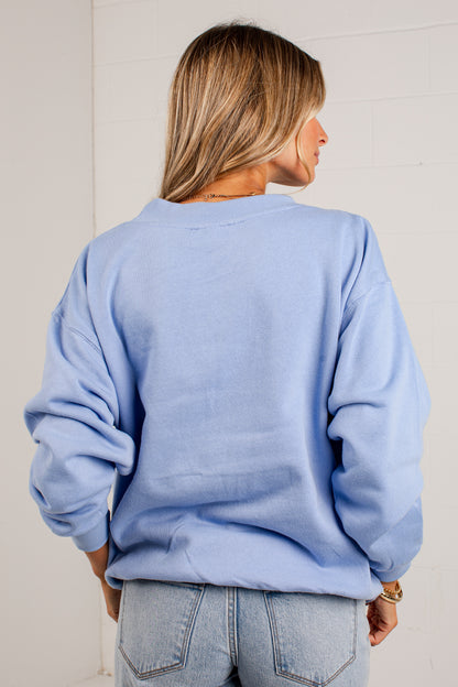 A woman viewed from behind wearing an oversized light blue crewneck sweatshirt and light-wash jeans against a white paneled wall.