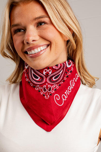 A smiling blonde woman wearing a red bandana-style neck gaiter with a white paisley pattern and the word &