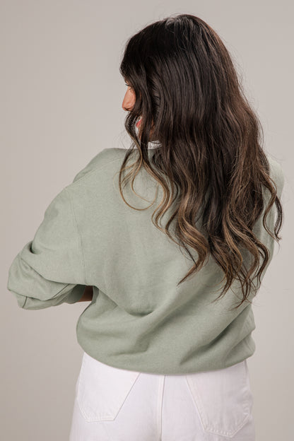 Woman wearing a light green sweater and white pants against a plain background