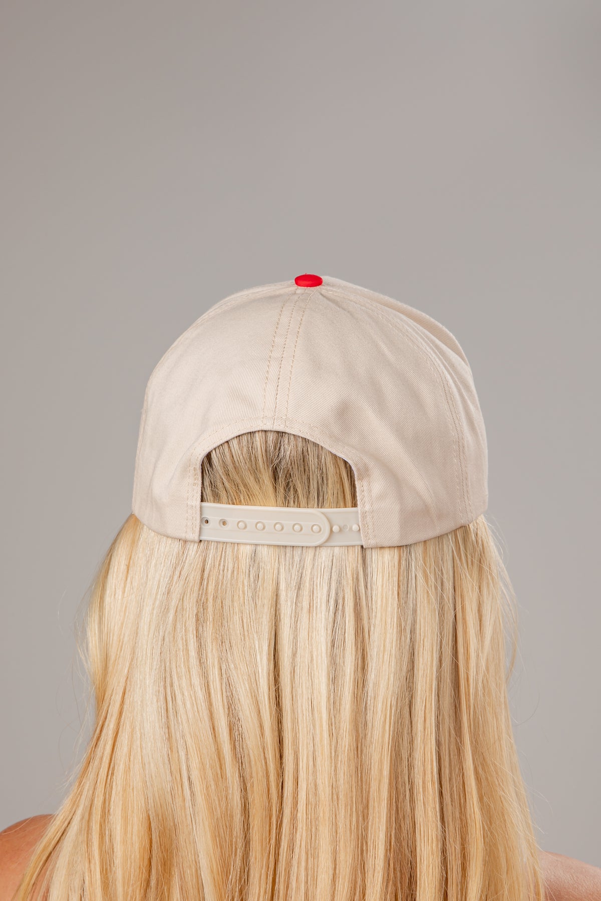 Beige cap with a red button on the back, worn by a person with blonde hair against a gray background