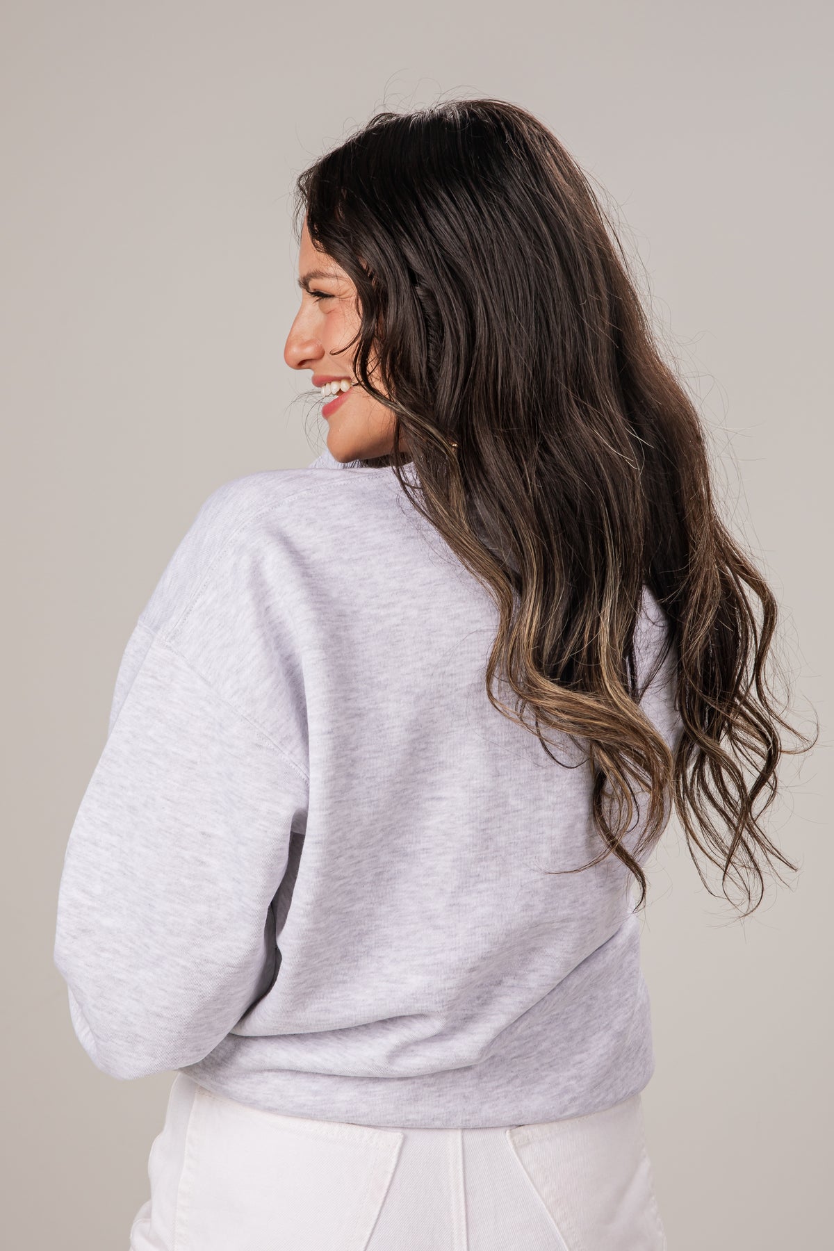 Woman wearing a light gray sweater and white pants against a plain background