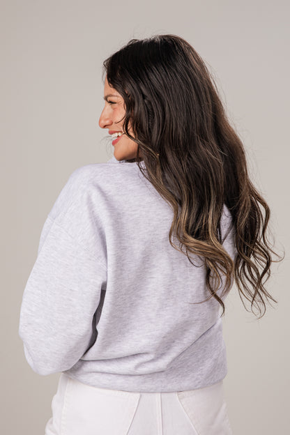 Woman wearing a light gray sweater and white pants against a plain background