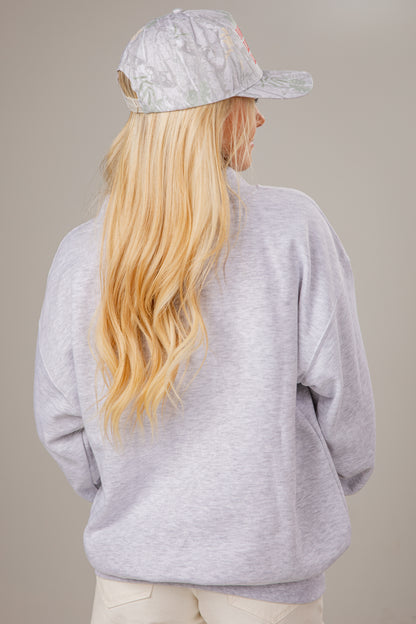 Person wearing a gray mocneck sweatshirt and cap with long blonde hair on a beige background